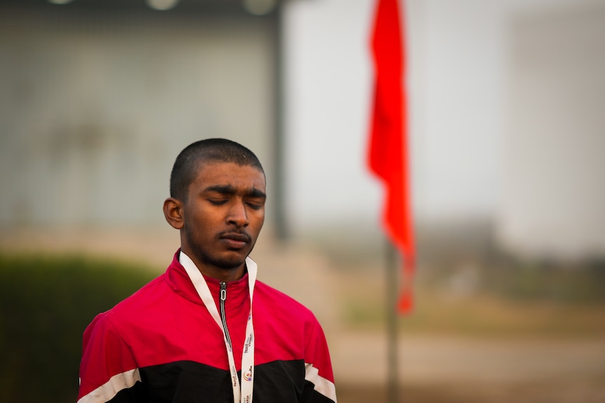 A man with his eyes closed stands near an orange flag dangling from a pole
