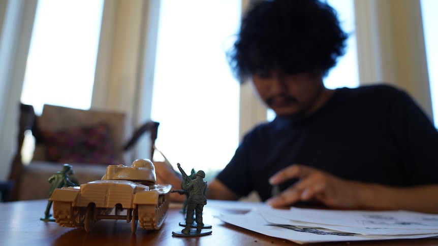 A young man seated on the floor leans on his arma s his draws on paper. Toy soldiers and tank sit on the table 