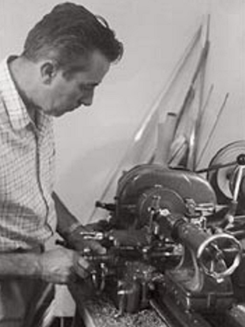 Black and white image of a man in chequered shirt bent down over an old fashioned sewing machine.