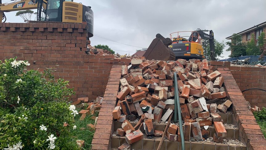 A pile of bricks tumble down a set of stairs in front of a demolition digger.