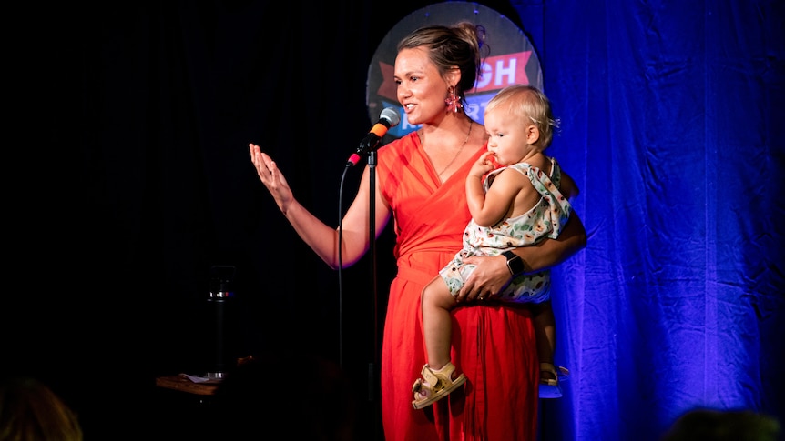 A blonde woman in a red dress performing on stage in front of a mic with a baby on her hip