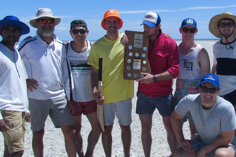 A group of men with a cricket bat and a wooden trophy standing on the beach.