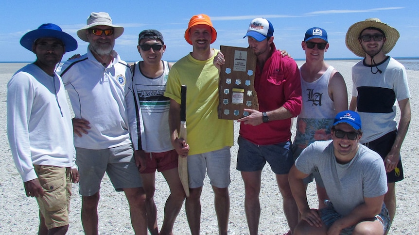 A group of men with a cricket bat and a wooden trophy standing on the beach.