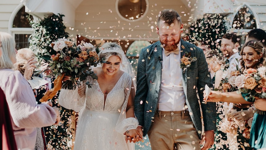 A newly married couple hold hands at their wedding as rose petals are showered over them by guests