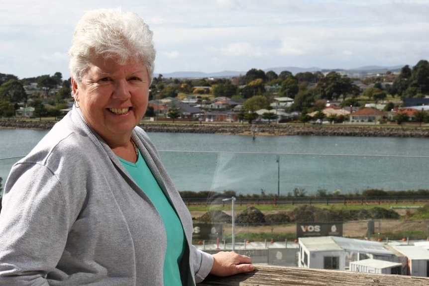 A woman with white hair stand on a deck, overlooking a river.