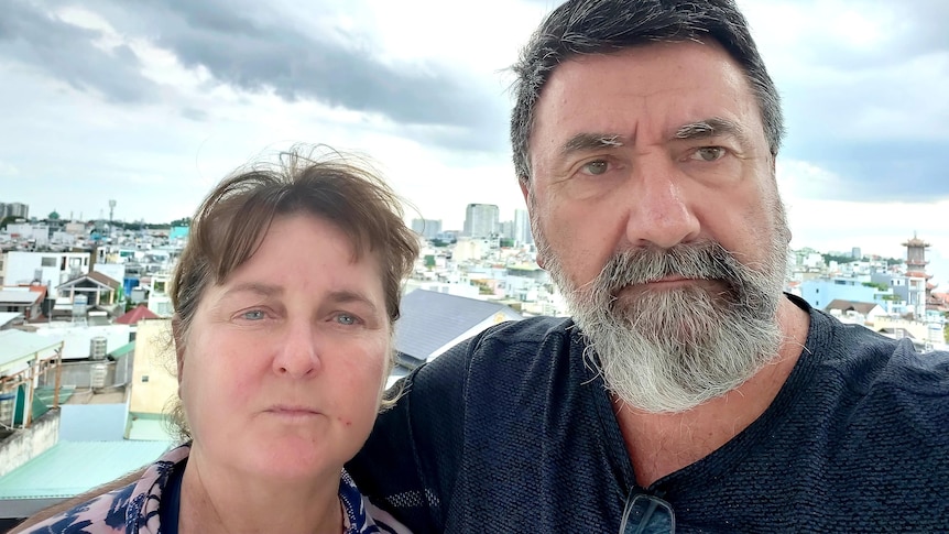 A middle-aged couple in a balcony selfie with a South-East Asian city sprawling out behind them.