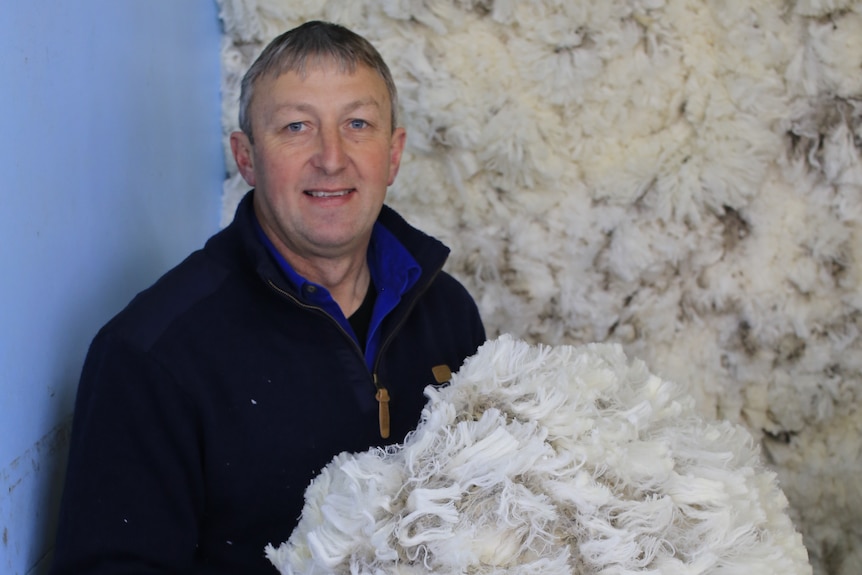 Trevor Mibus holds up a pile of Merino wool. He stands in front of more Merino wool.