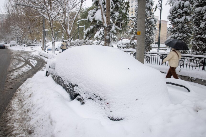 人们路过停在街上的一辆积雪覆盖的汽车。