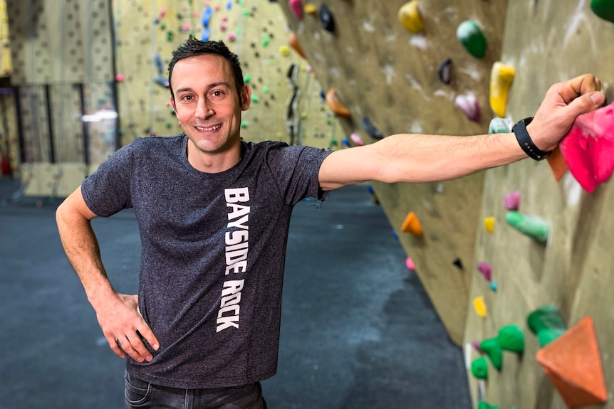 A man smiles for a portrait while leaning on a rock climbing wall