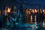 A male and a pregnant female Na'vi stand in water surrounded by other Na'vi holding torches