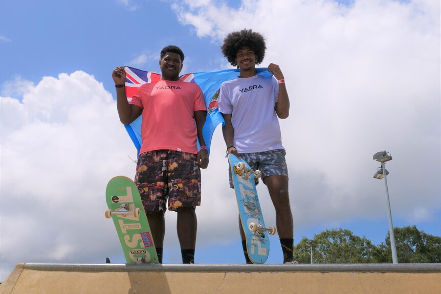 Two Fijian boys stand at the top of a skate bowl holding their country flag.