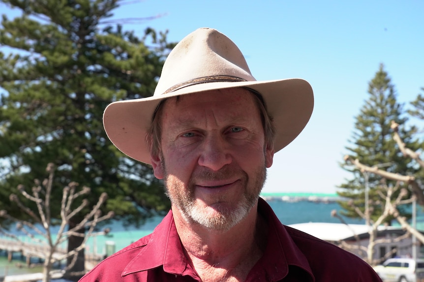 A close up of a man wearing a tan coloured hat smiling at the camera with water and trees in the background