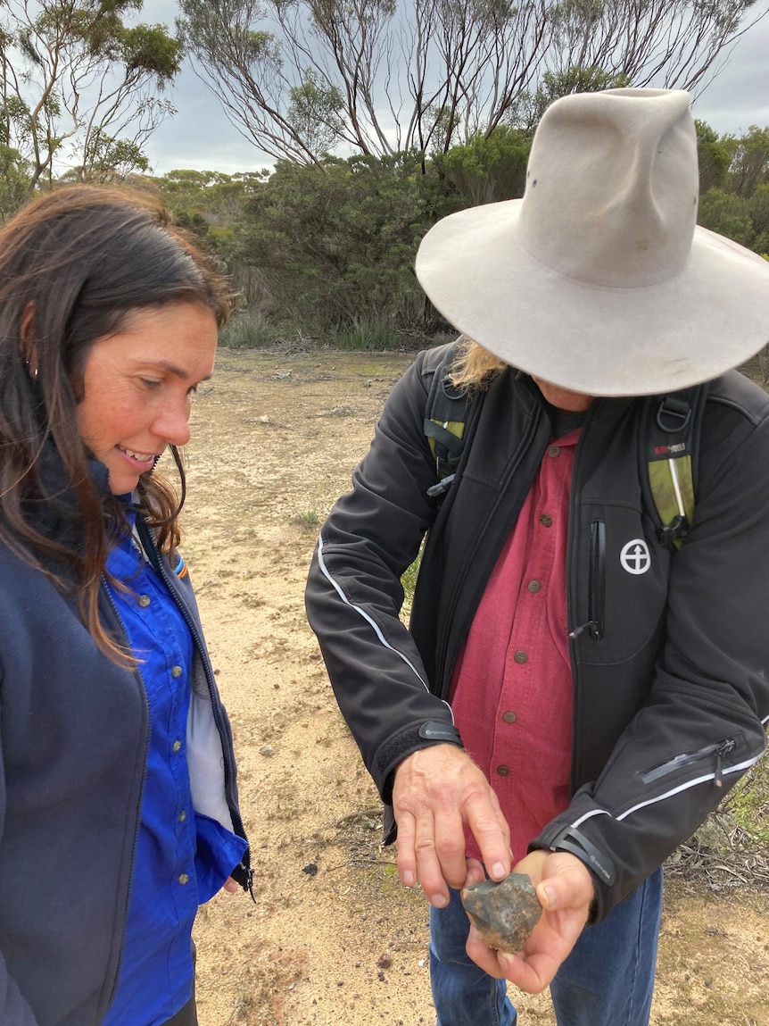 A woman in a blue jumper is standing next to a man in jeans and a brown hat looking at an Aboriginal artefact in his hand.