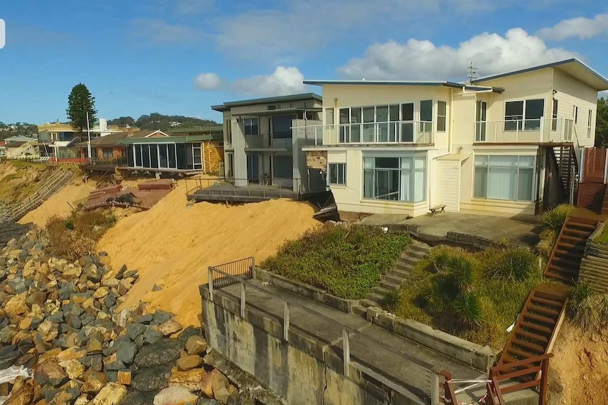 A drone photo of two houses near the ocean with sand and rock pushed in front of them