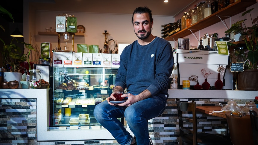 A man sits on a stool in the middle of a cafe. He has a cup of coffee in his hand.