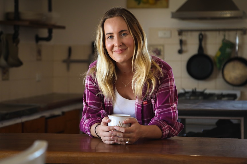A young blonde woman stands in a rustic kitchen. She's smiling and holding a coffee mug.