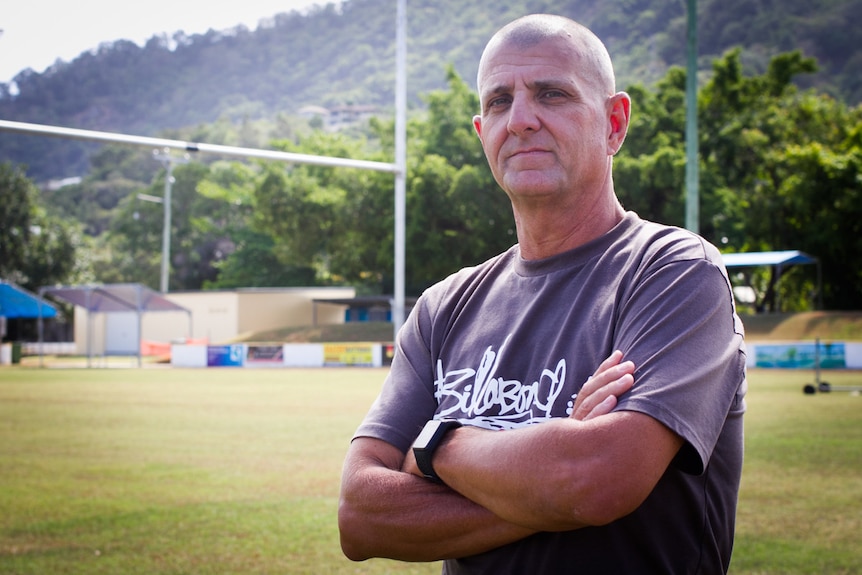 A bald man standing on a sports oval with goalposts in the background.