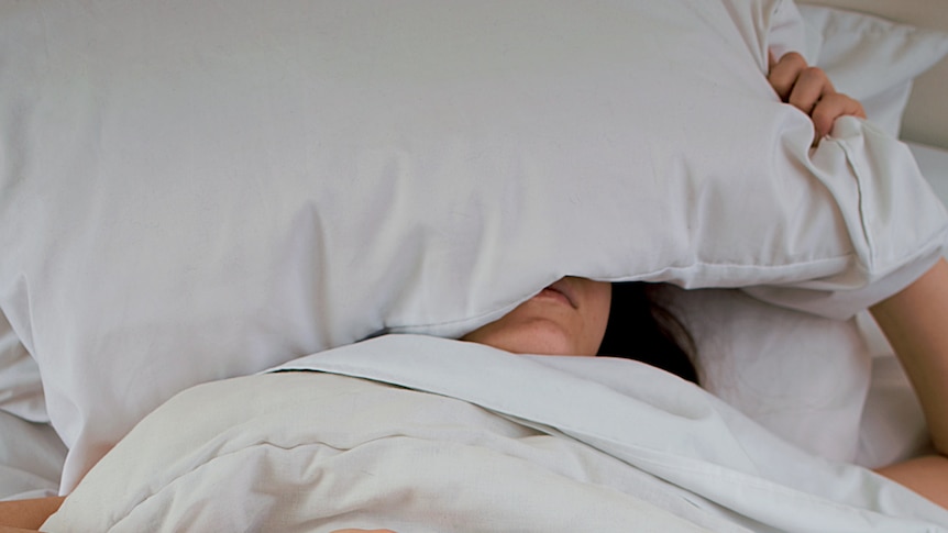 Woman with a pillow over her head and holding glasses in her hand for a story about the best time to go to sleep.