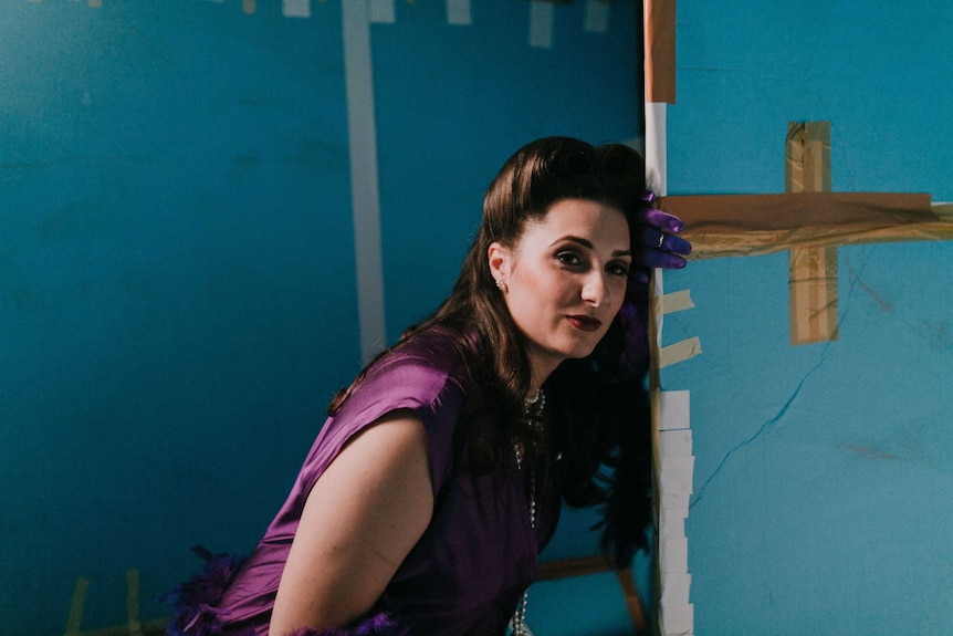 A brunette woman in a fancy purple dress leaning against a patched up blue wall