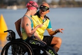 woman in in green and gold swim gear sits in wheelchair next to bald man talking to her