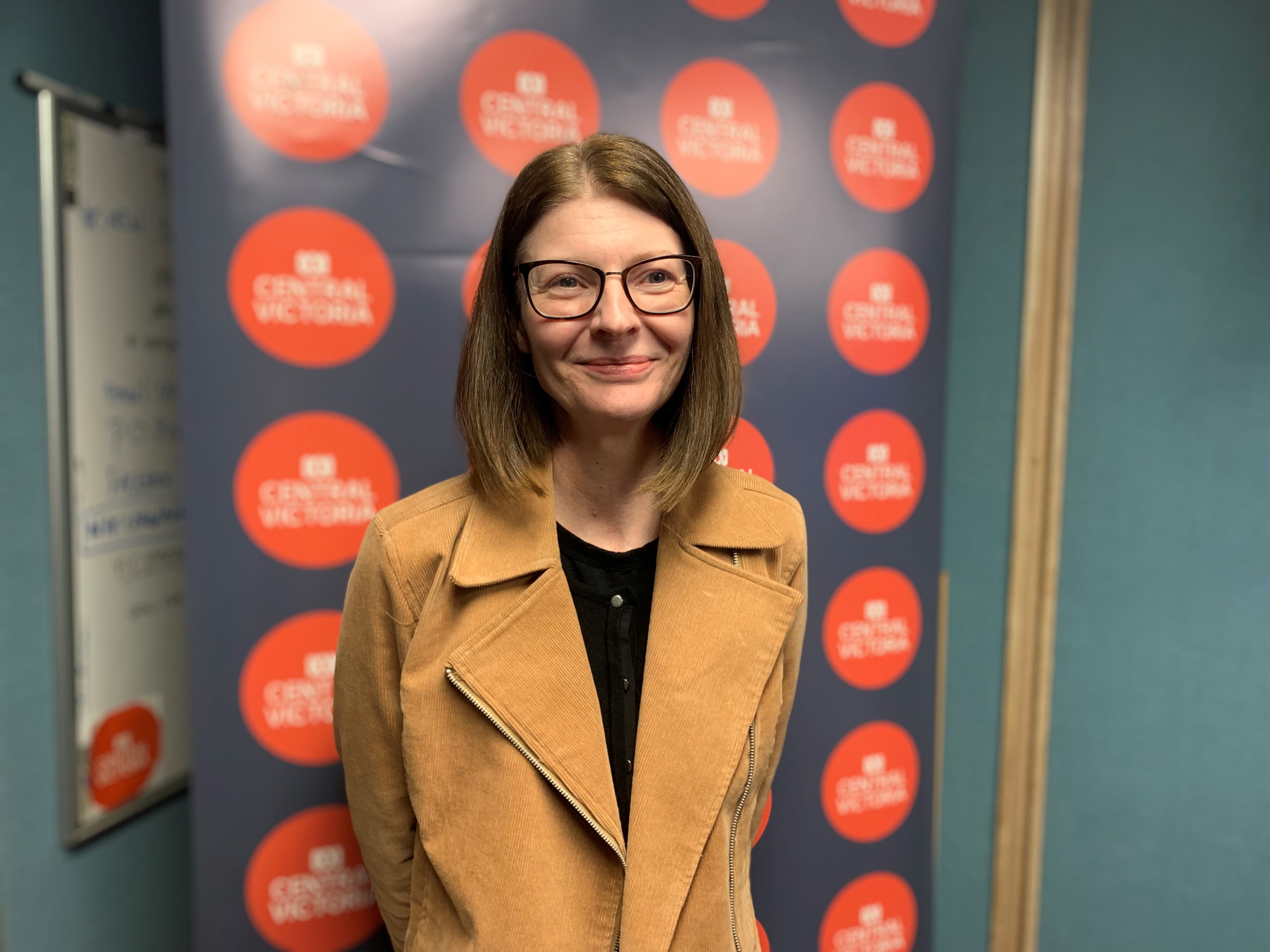 A woman with brown shoulder length hair, wearing glasses and a tan jacket.