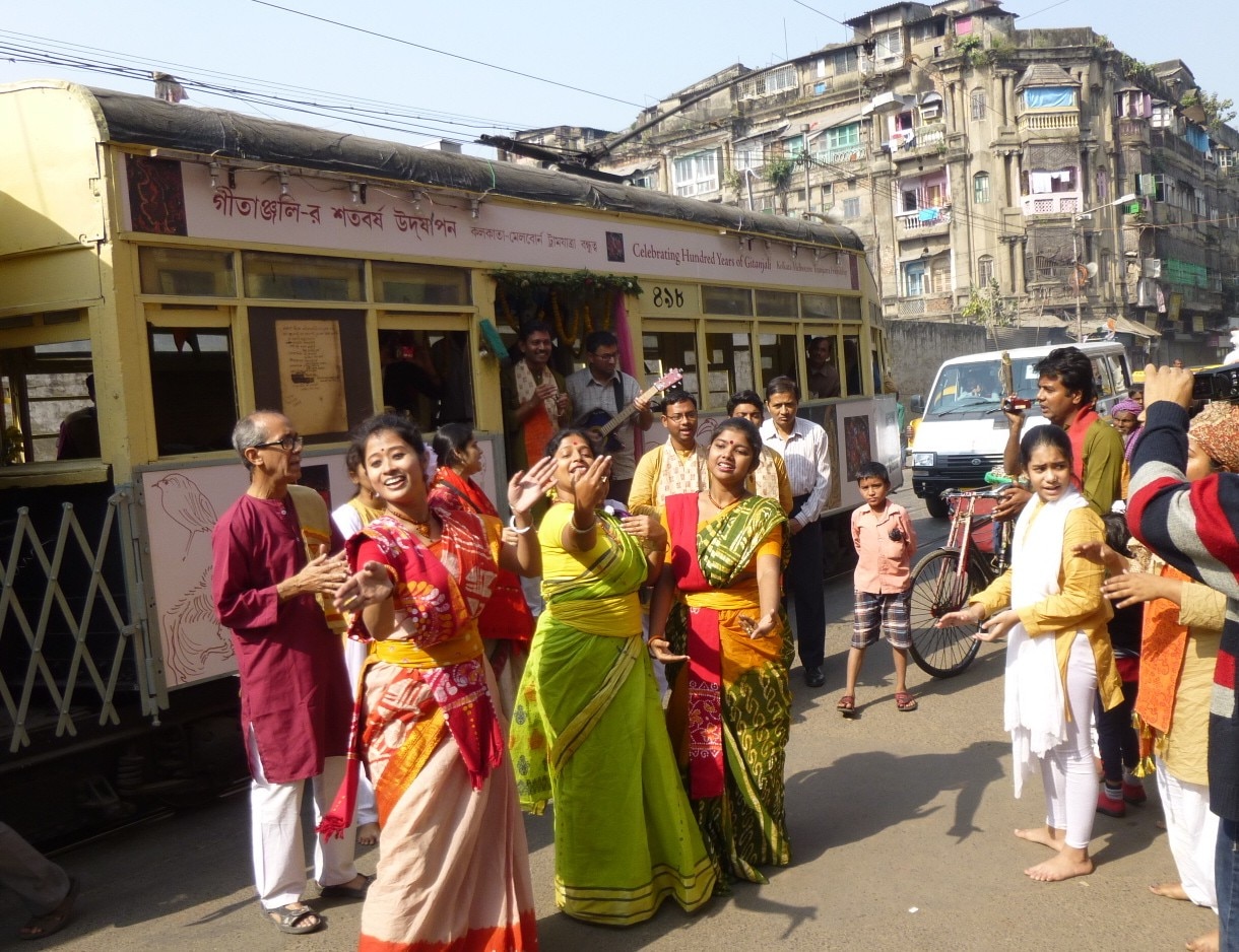 Un grupo de mujeres con saris bailan frente a un tranvía en una calle india.