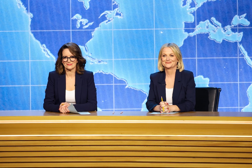 Tina Fey and Amy Poehler sit at a pretend news desk in news anchor suits.