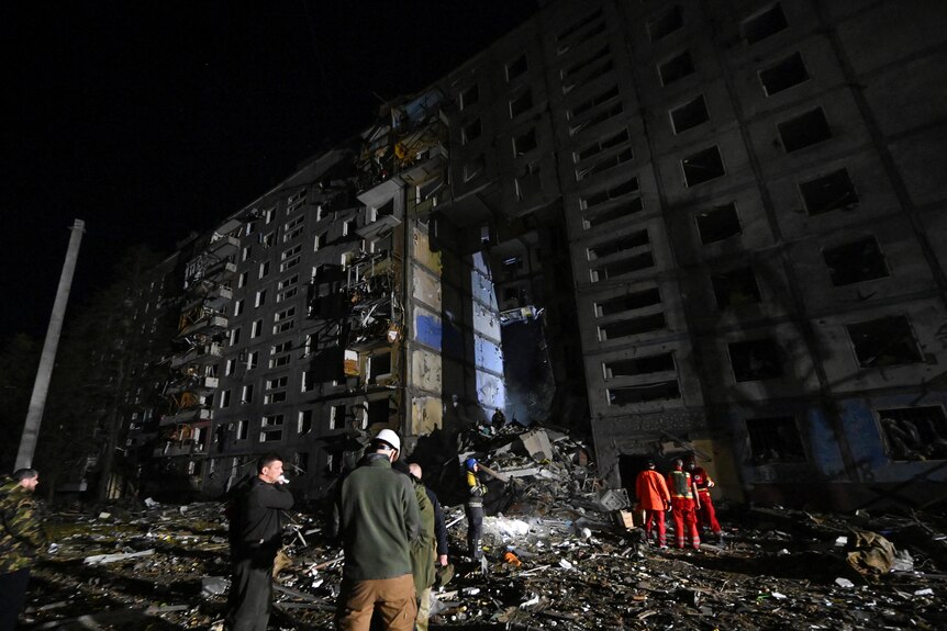 rescue workers stand in rubble next to a damaged residential building at night