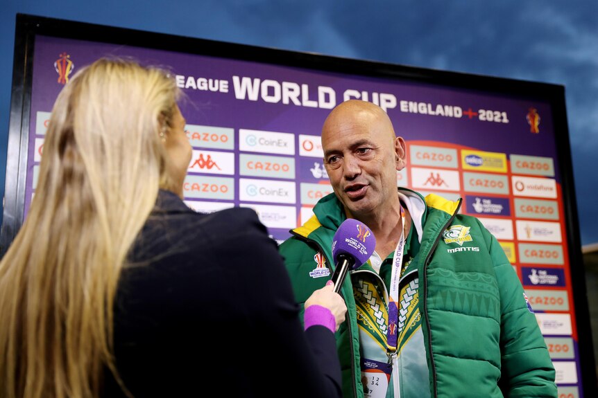 A rugby league coach speaks to a reporter holding a microphone at the ground before a game.
