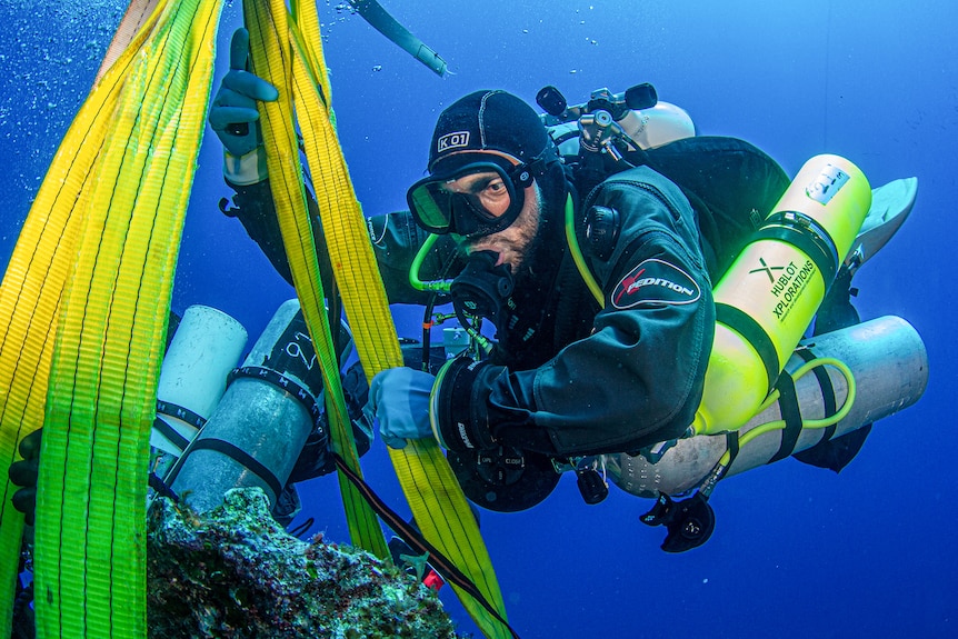 A diver wearing a wetsuit and holding oxygen tanks helps guide a boulder being lifted by a yellow rope.