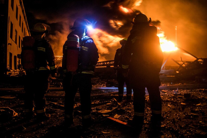 The silhouettes of four firefighters, standing facing a roaring fire and billowing smoke, are visible in the foreground.