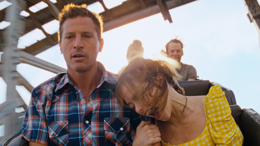 A young woman holds the arm of a frightened-looking middle-aged man as they ride a rollercoaster