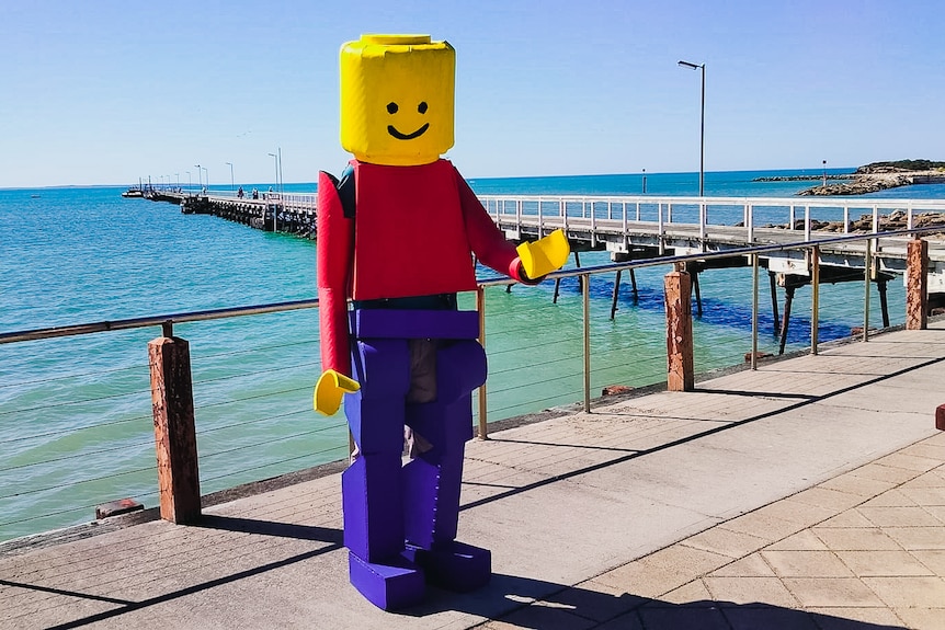 A man in a blue and red foam Lego suit stands near a jetty, by crystal clear sea waters on a sunny day.