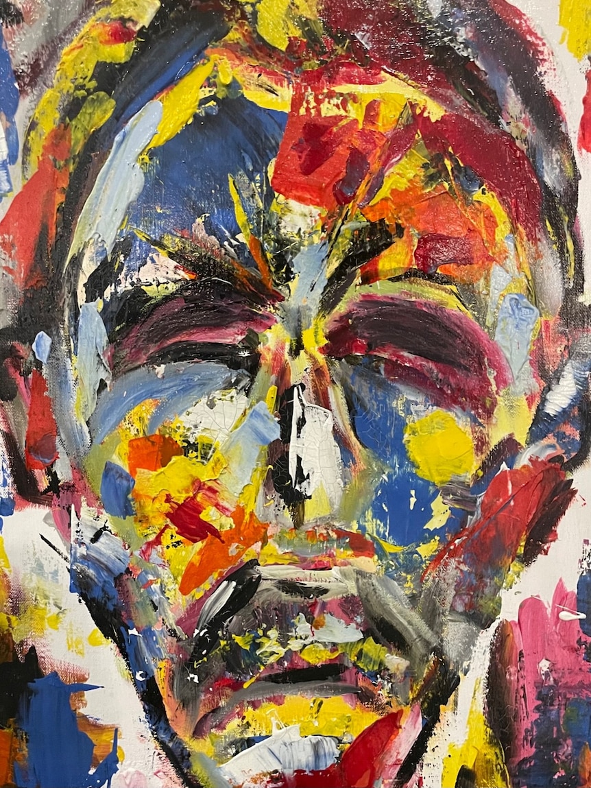 An abstract painting of a somber man's face in bright yellow, red, blue, black and grey  