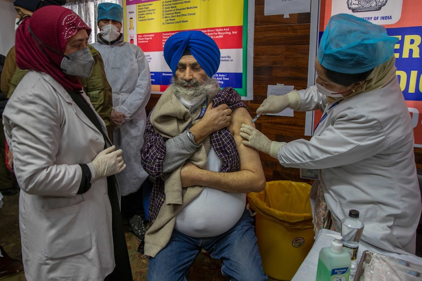 A nurse in a white robe vaccinates a man in a turban.