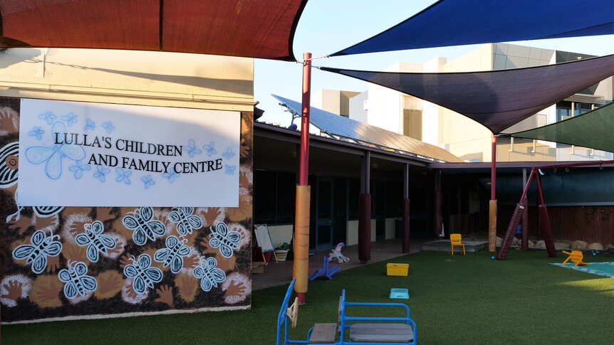 The front yard of a daycare centre with toys and chairs on an artificial lawn and a sign on the wall naming the business.