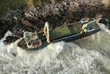 An unmanned cargo ship is wedged against a rugged coastline as waves push it against the rocks