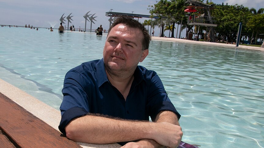 A man with short dark hair, wearing a navy shirt, sitting in a pool.