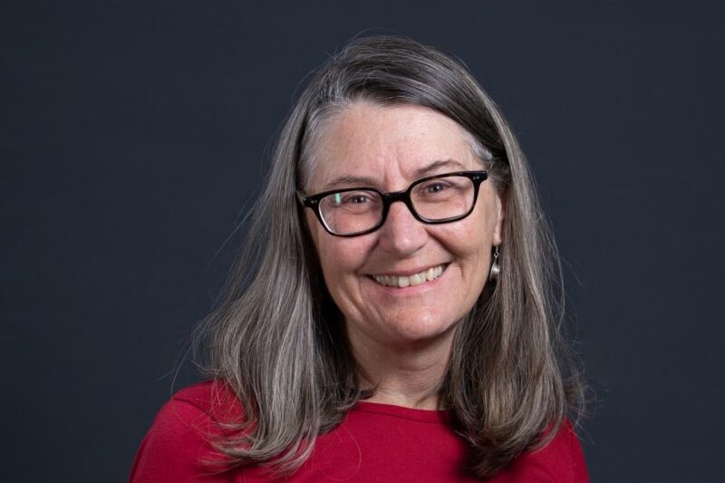 A headshot of Doctor Andrea Sharam, she is wearing glasses with long hair and a red shirt, smiling with grey background
