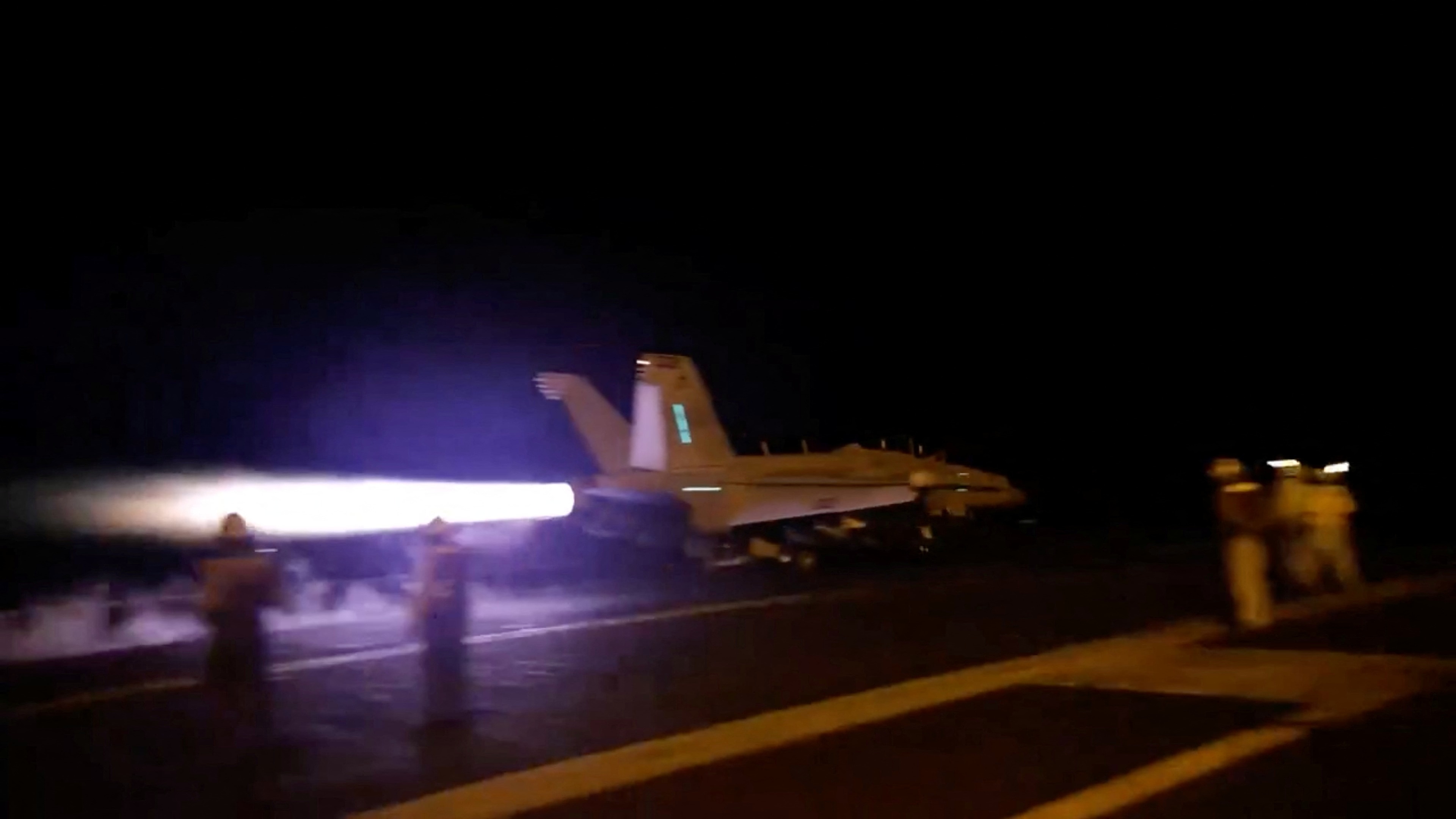 A jet aircraft shoots across the flight deck of an aircraft carrier at night.