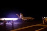 A jet aircraft shoots across the flight deck of an aircraft carrier at night.