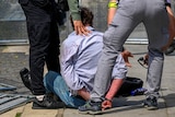 A man sitting on the ground with handcuffs on, with his back to the camera, surrounded by other men.