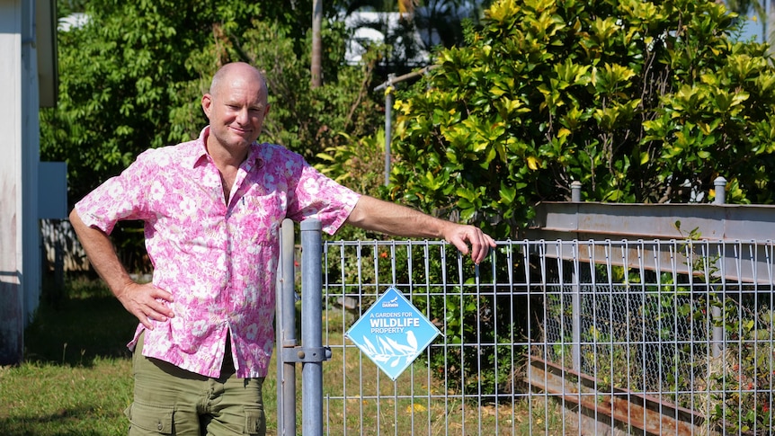 Man in pink floral shirt leaning against a wire fence carrying a "gardens for wildlife property" sign