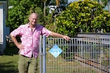Man in pink floral shirt leaning against a wire fence carrying a "gardens for wildlife property" sign
