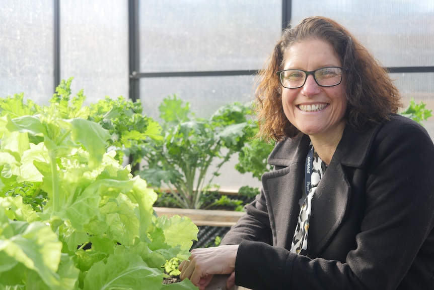A woman with brown hair and glasses smiles at the camera from within a greenhouse. 