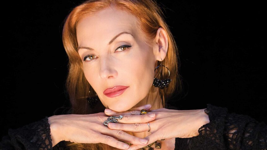 A portrait of Ute Lemper with her hands folded under her chin in front of black backdrop.