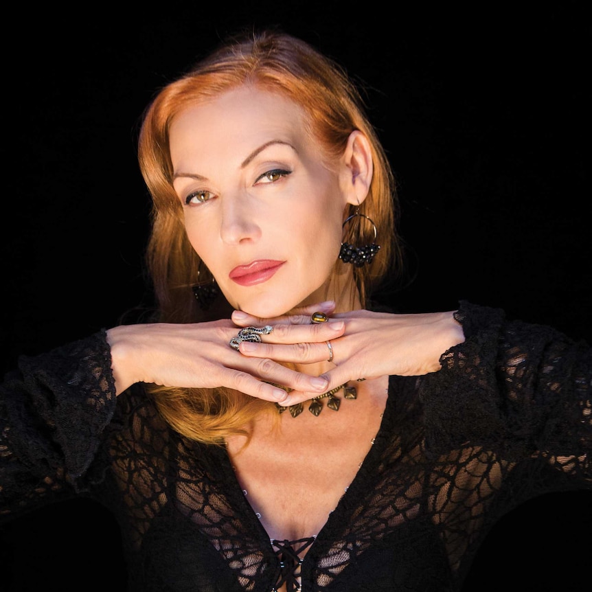 A portrait of Ute Lemper with her hands folded under her chin in front of black backdrop.