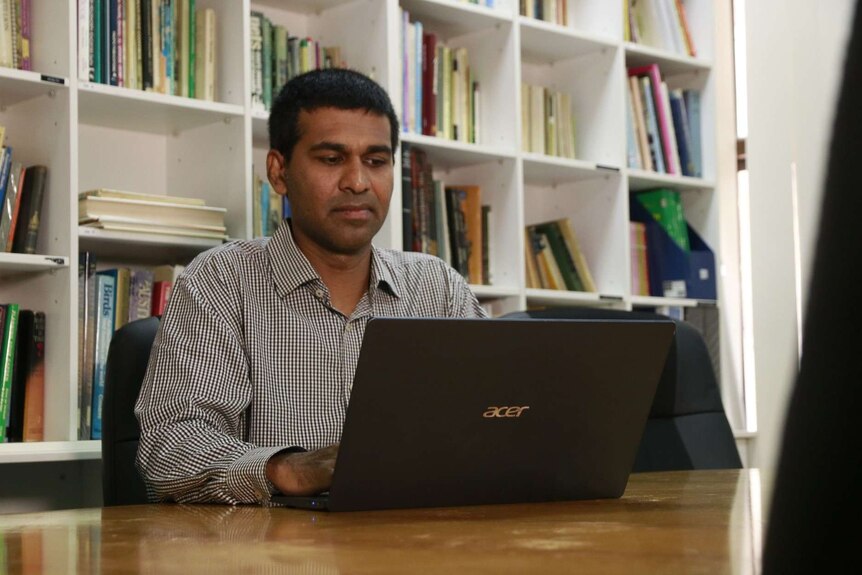 A photo of Edwin Joseph sitting at his computer with a bookshelf behind him.