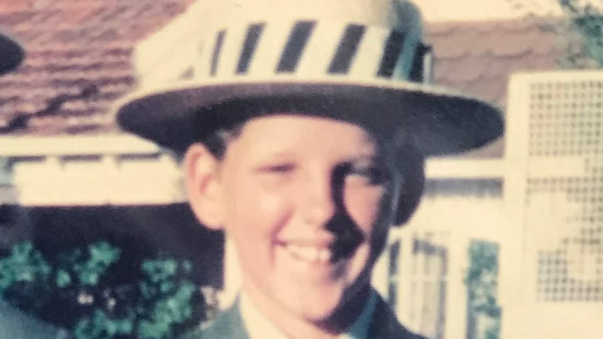 A boy stands smiling in a grey suit with a straw hat with a black and white striped band