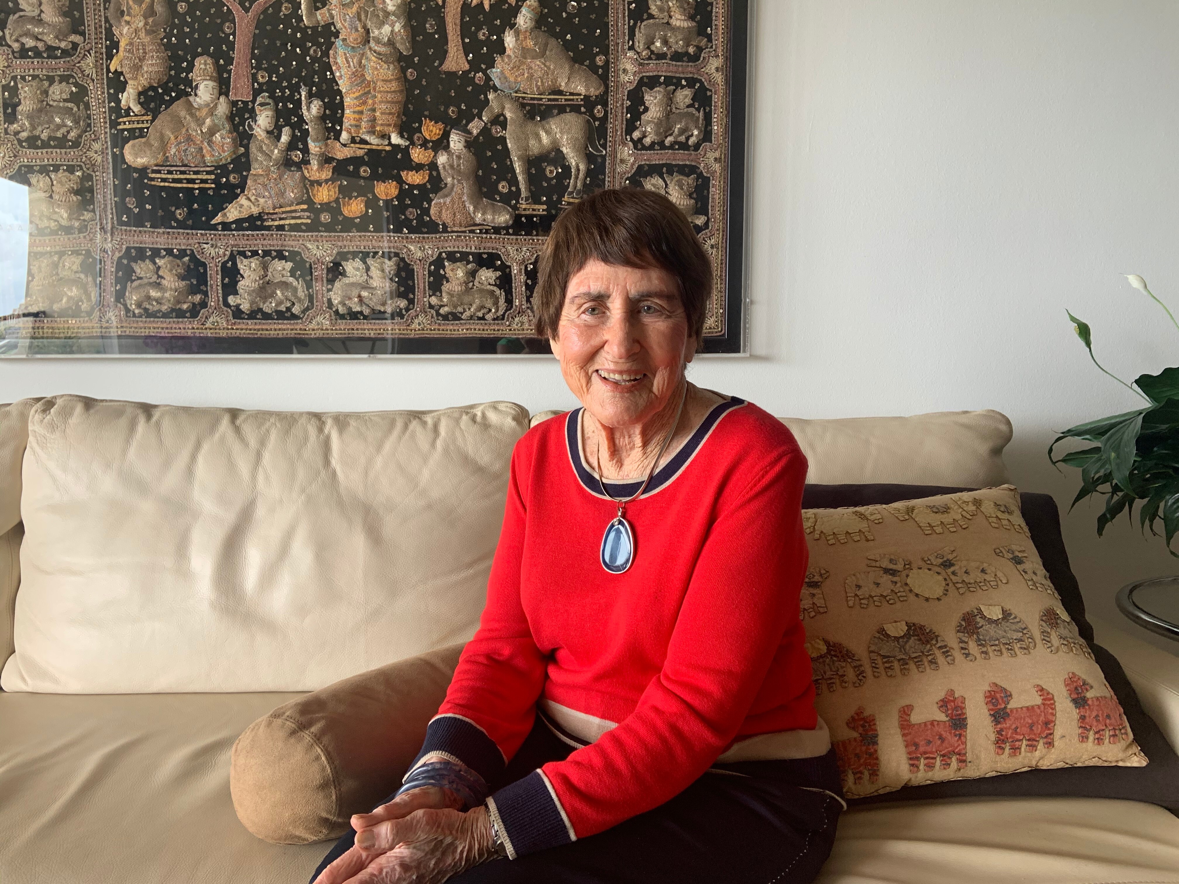A woman in a red jumper with dark brown hair sits on a cream couch with a Thai tapestry on the wall behind her.